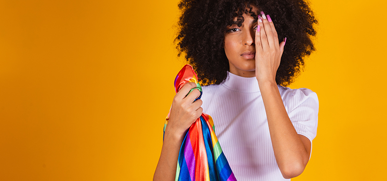 Mulher negra com cabelos crespos com a mão em frente ao olho direito, segurando a bandeira do orgulho LGBTQIAPN+. Foto: Adobe Stock