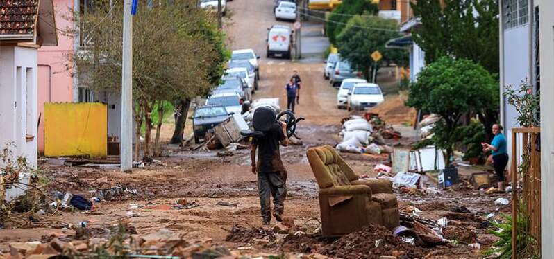 Pessoa levando uma cadeira em meio a rua com objetos do desastre no Rio Grande do Sul em volta. Foto: Divulgação/ Gustavo Mansur