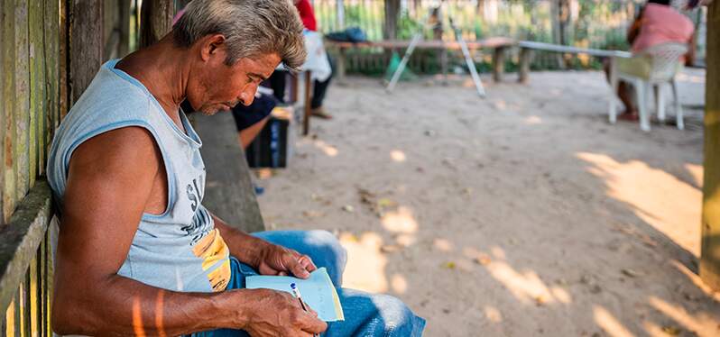 Na imagem, um homem, de regata e bermuda, aparece sentado, segurando caneta e folhas em suas mãos.