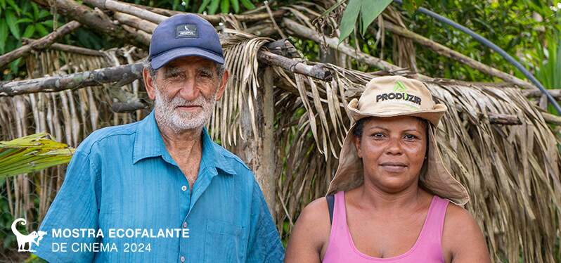 Na imagem, é mostrado um casal que reside na região do Médio Xingu. O homem utiliza camisa e boné azuis, enquanto a mulher veste uma regata rosa e um chapéu. Atrás deles, uma casa feita de madeira.