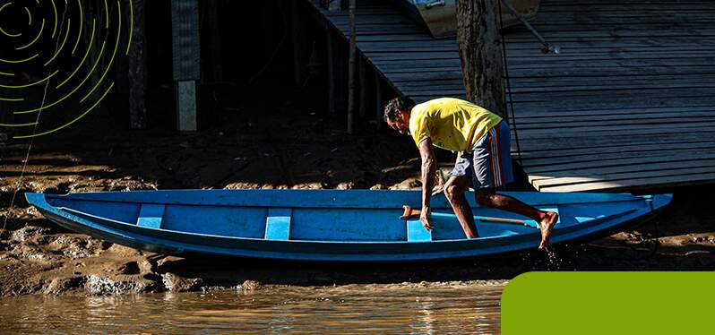 Na imagem, um homem com camiseta amarela está à beira de um rio, em cima de um barco azul.