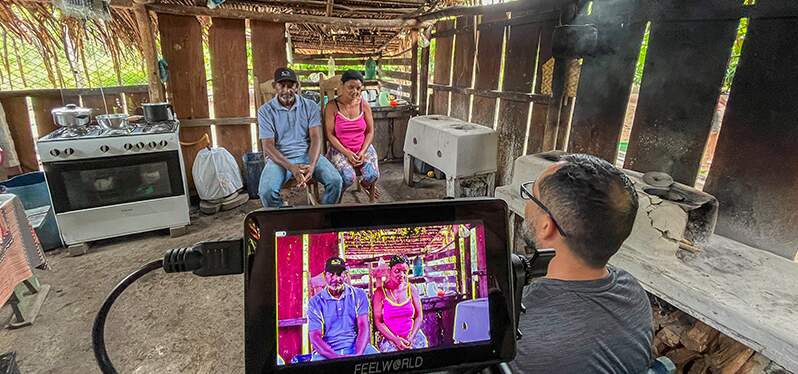 Na imagem, um casal heterossexual está sentado, em cadeiras, dentro de uma casa, sendo filmado por uma câmera de vídeo