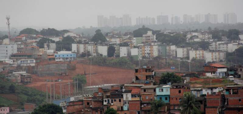 A imagem mostra um retrato da desigualdade social, onde aparecem na frente uma comunidade e uma conjunto habitacional, enquanto bem no fundo da imagem são mostrados grandes prédios